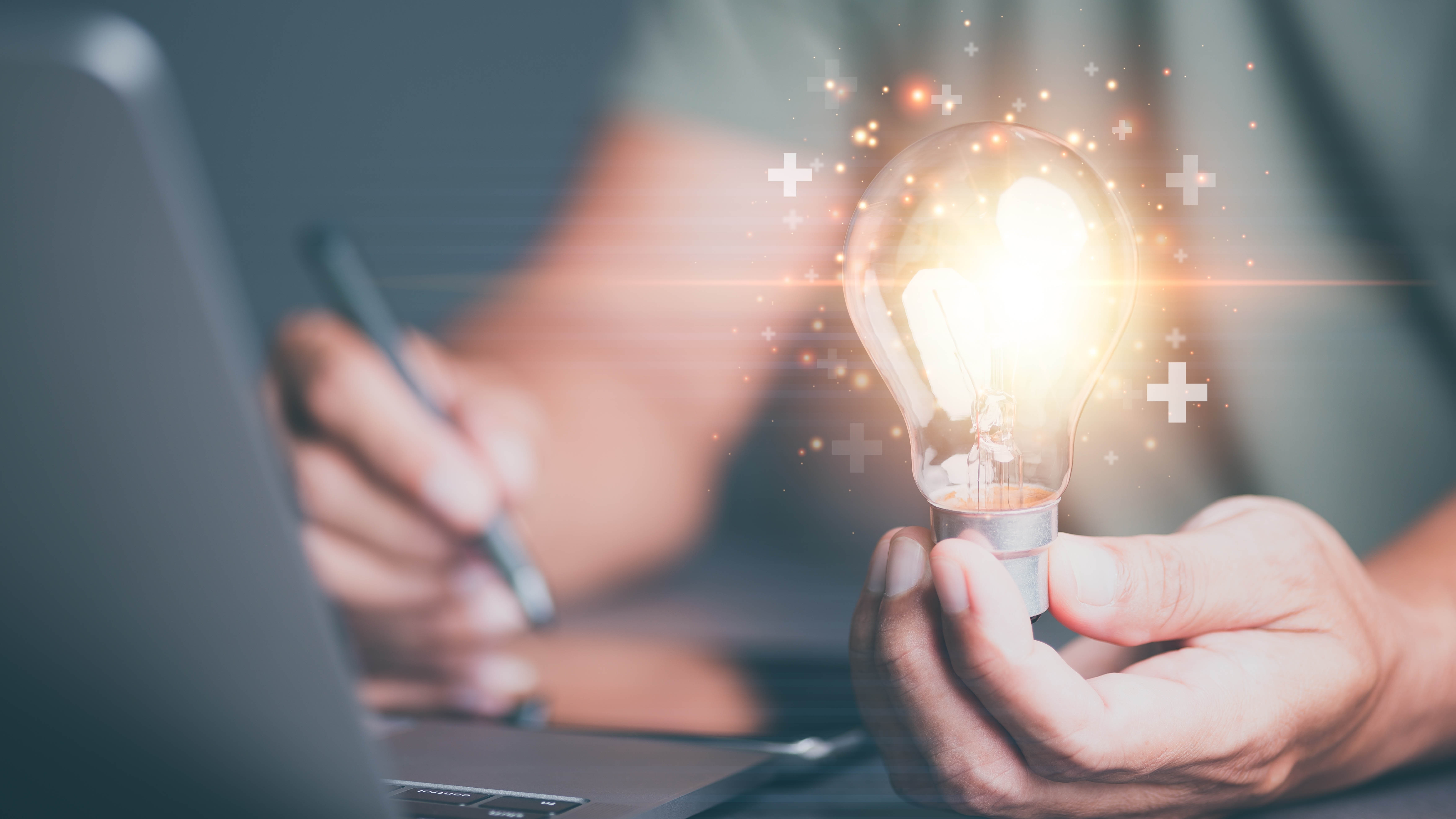 Close-up of hand holding a lit lightbulb in front of a computer